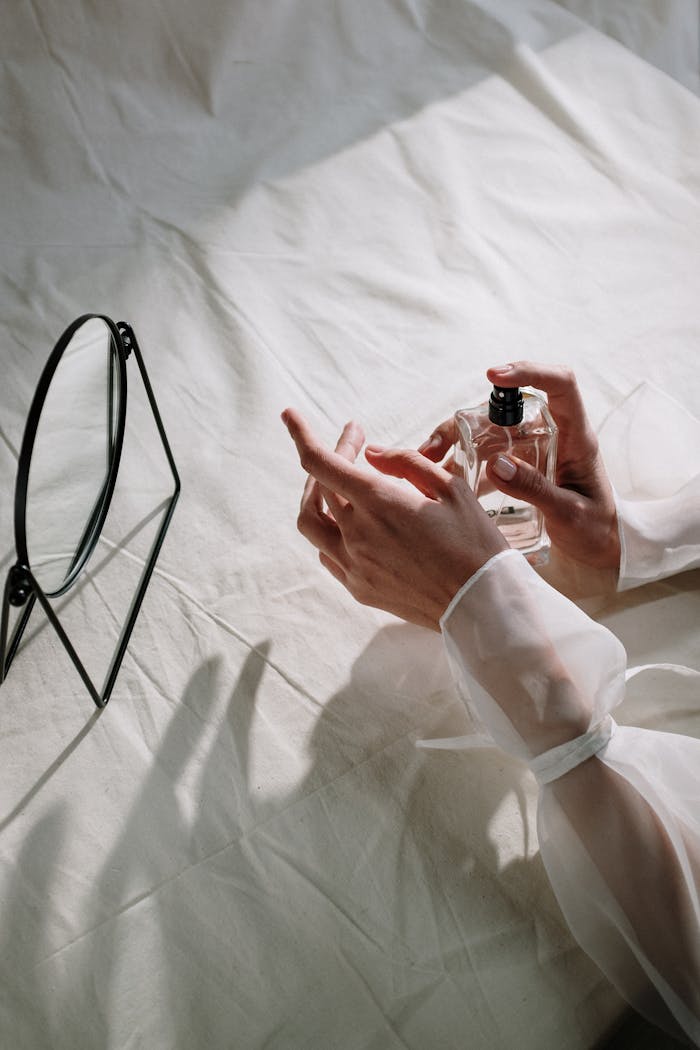 creative-02 Delicate hands holding a perfume bottle beside a mirror on a white cloth background.